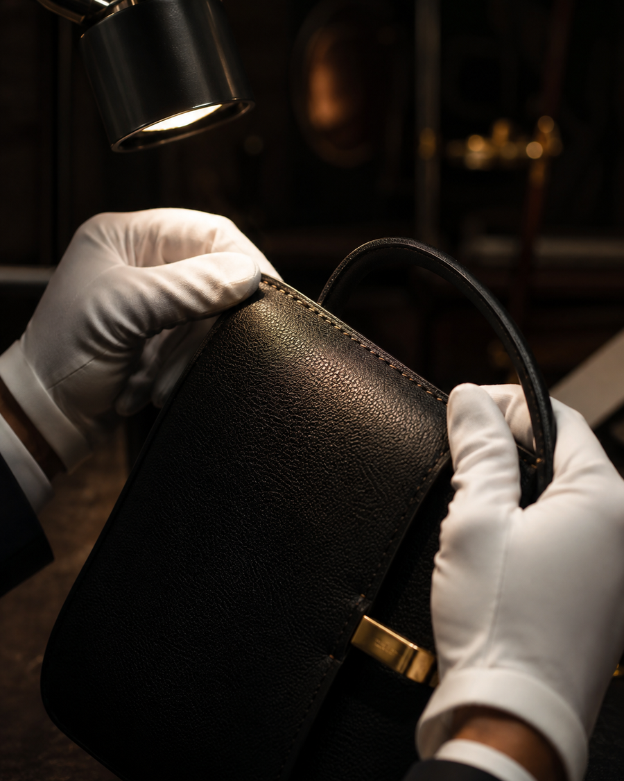 White-gloved hands inspecting a black leather handbag under a jeweler's lamp